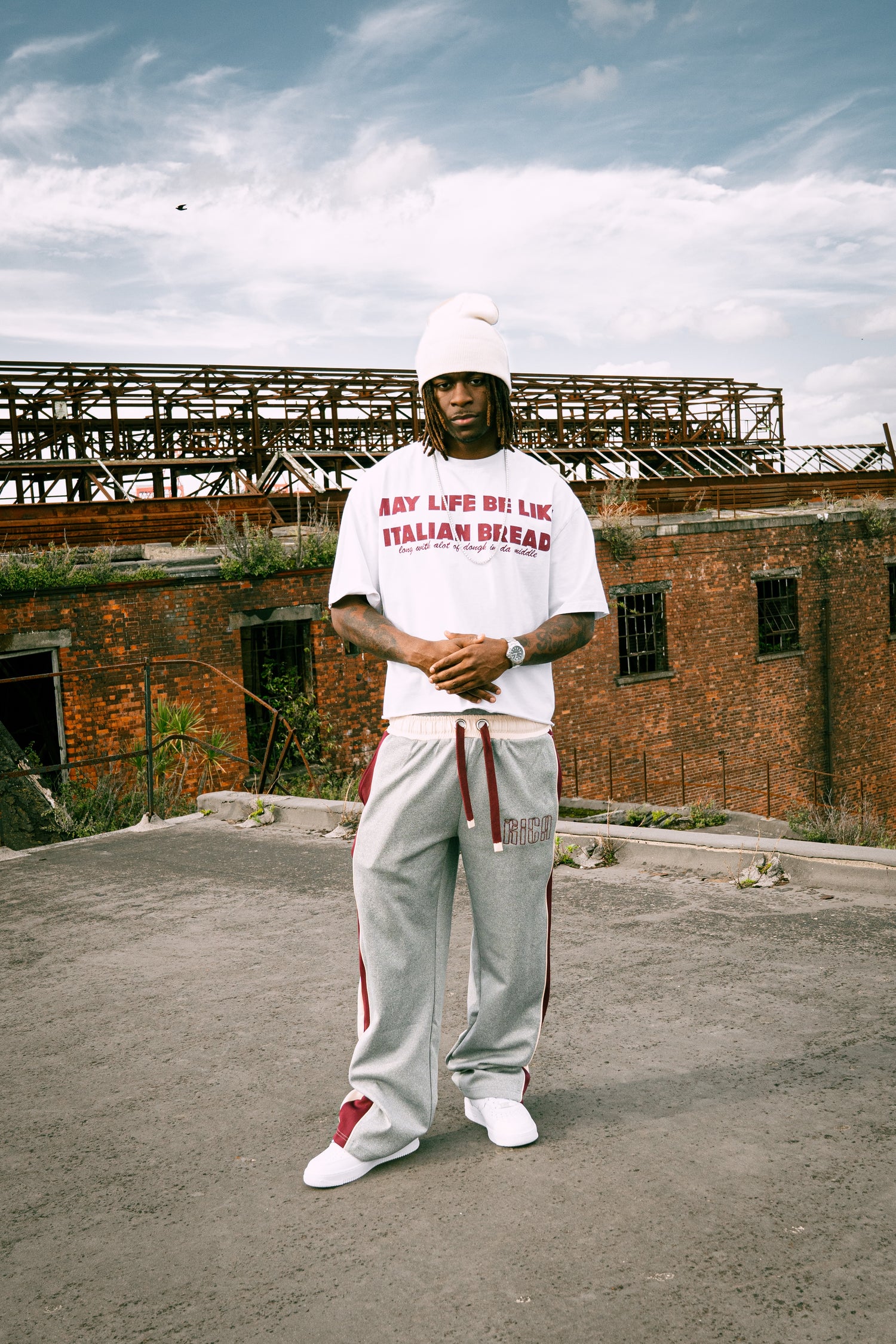 Person wearing a white t-shirt with burgundy text saying "may life be like italian bread" and gray, cream and burgundy pants standing in front of an abandoned building. RICA STILLRICA 